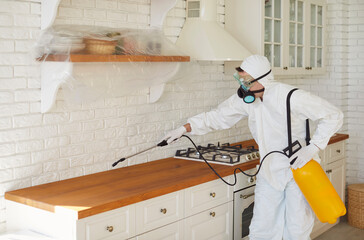 Professional exterminator wearing protective suit, gloves and respirator mask disinfecting modern kitchen. Pest control specialist using sprayer with chemical solution for pest control or sanitation.