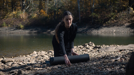Woman rolling out yoga mat by a rocky lakeshore in the forest
