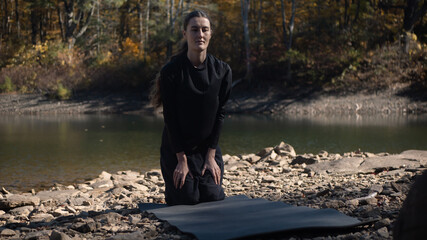 Woman kneeling on yoga mat by rocky riverbank in autumn forest
