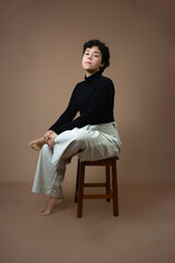 Young woman with short curly hair, sitting on a stool with crossed legs and hand on ankle. Vertical studio shot with copy space, perfect for Gen Z, cool attitude, female empowerment and femininity.