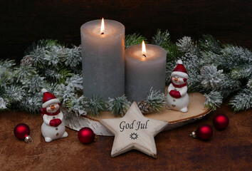 Gray burning candles are arranged on a wooden disc, decorated with Christmas baubles, fir branches, and decorative figures. The text "God jul." is written on a star.