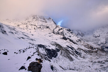 A snowy mountain peak rises into a cloudy sky. The location is the Annapurna Sanctuary, a popular destination in the Nepal Himalayas for hikers