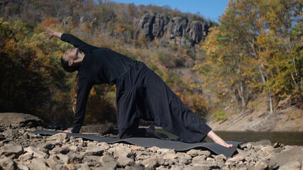 Woman practicing yoga pose outdoors on rocky riverbank in autumn