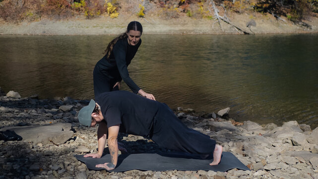 Outdoor yoga session with instructor helping student by riverside - Powered by Adobe