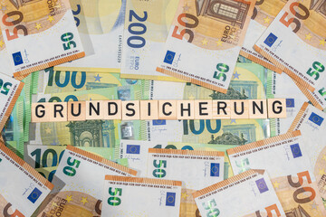 Wooden tiles form Grundsicherung on a spread of euro banknotes. Concept image for social security,...