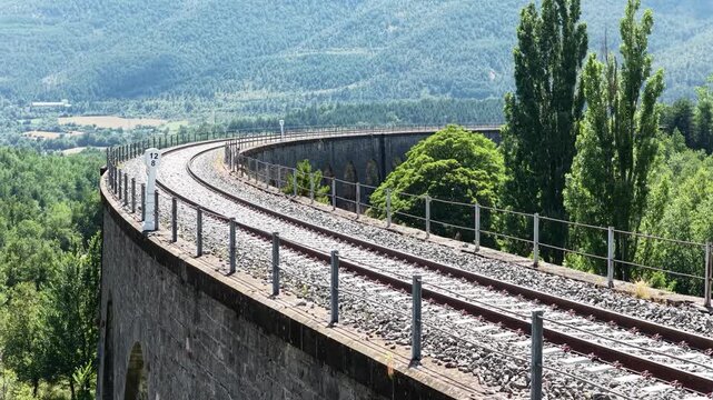 Train tracks curve gracefully on a viaduct, set against a backdrop of lush green mountains and valleys