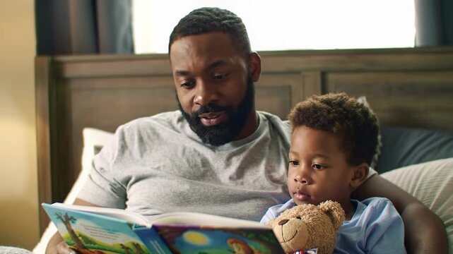 Warm, intimate video: African American father (mid-30s) reads vivid storybook to his son (5) on a plush bed with golden light and bokeh. Concept of family bonding and warmth