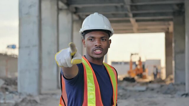 Confident african american male builder in a hard hat pointing at the viewer on a construction site. Professional foreman or engineer giving instructions and managing the project