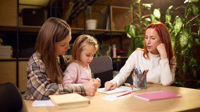 Family scene of two mothers assisting little girl with homework. Concept of child development, supportive study environment, inclusive family values, and modern learning culture.