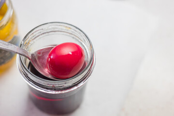 A close-up of the process of dyeing an Easter egg. The egg is being removed from the dye in a jar with a spoon. Other jars of dye are visible in the background