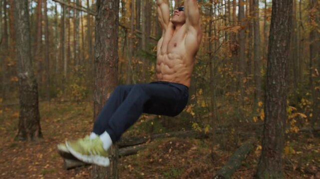 Young muscular man training on makeshift sports ground in autumn forest and performs leg raises on horizontal bar nailed to tree.