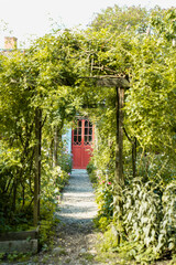 ein Kiesweg durch einen verwachsenen Garten mit einer roten T&uuml;r am Ende des Weges