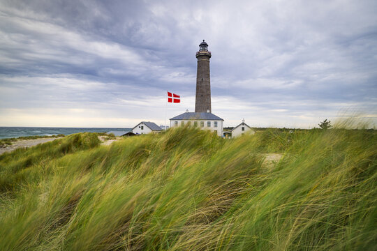 View of a towering lighthouse with a Danish flag fluttering against a backdrop of windswept dunes and a moody sky, Skagen, Denmark.