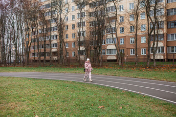 78-year-old senior woman walking with nordic poles at stadium. Pensioner practice nordic walk in city. Healthy retiree lifestyle, outdoor senior fitness.