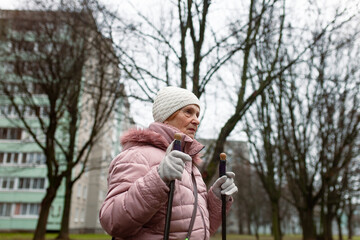 Close up of senior woman walking with nordic poles in a city. 78-year-old pensioner practice nordic walk near residential buildings. Healthy retiree lifestyle, outdoor senior  fitness.