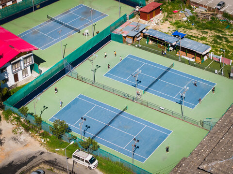Aerial view of tennis courts alive with players under the open sky, framed by traditional Bhutanese architecture and lush greenery, Thimphu, Bhutan.