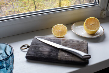 Knife on dark wooden cutting board with lemon halves on windowsill. Kitchen prep, fresh ingredients, natural light. Real photo