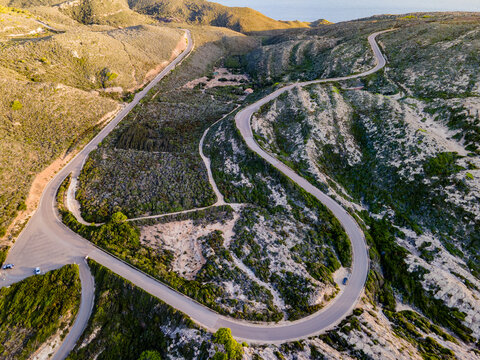 Aerial view of a winding road snakes through sun-drenched hills, where shadow and light dance across the landscape, revealing rugged terrain, Capo Sandalo, San Pietro, Sardinia, Italy