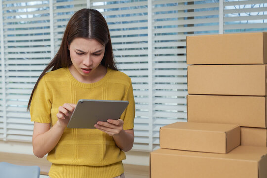 Asian businesswoman confused reading tablet data surrounded by boxes showing frustration and stress due to e commerce management issue mistake or system error in digital business.