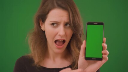 A surprised young woman with a shocked expression holds up a smartphone with a green screen for display against a green background. - Powered by Adobe