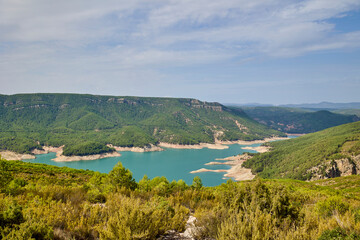Wide panoramic view of a turquoise mountain lake surrounded by green forests and hills in Spain under a partly cloudy sky. Peaceful natural landscape and wilderness scenery.