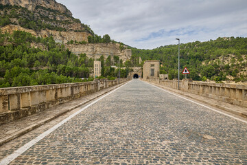 Scenic view of a cobblestone bridge road leading to an old stone tunnel surrounded by forested mountains under cloudy sky.
