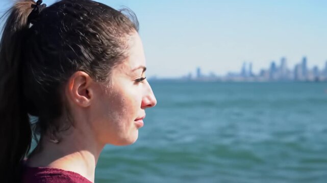 Close-up video of serene young caucasian woman with brunette ponytail gazing contemplatively at vast cerulean ocean, soft blurred city skyline, slow pan. Tranquil reflection concept