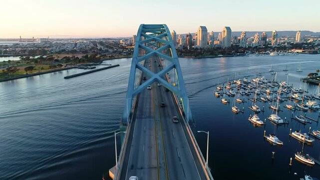 Sweeping aerial crane video dollying forward over iconic San Diego-Coronado Bridge at golden hour. Blue arching structure, tranquil sapphire bay with yachts, glowing city skyline. Serene urban