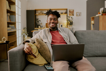 Portrait of young adult Black man sitting on sofa using laptop for online shopping, smiling at camera, holding credit card in hand, smartphone and cushions visible in foreground