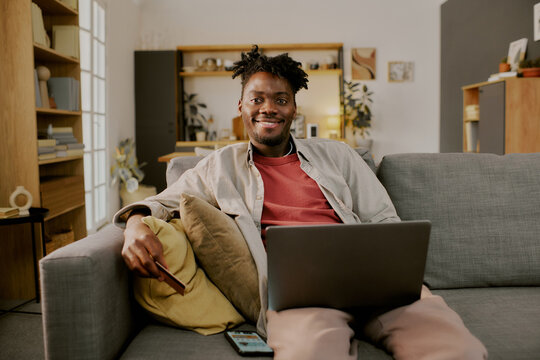 Portrait of young adult Black man sitting on sofa using laptop for online shopping, smiling at camera, holding credit card in hand, smartphone and cushions visible in foreground
