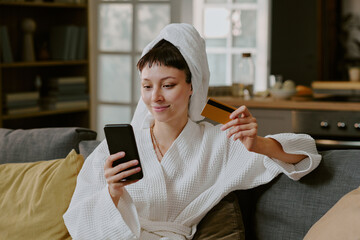 Young adult Caucasian woman sitting on sofa holding smartphone and credit card smiling while making online purchase at home, wearing bathrobe with towel wrapped around head