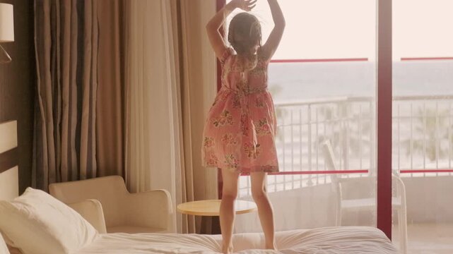 little girl in floral dress jumping on hotel bed, silhouetted against large window with ocean view, arms raised in pure joy and excitement, vacation fun, childhood joy captured in moment carefree play