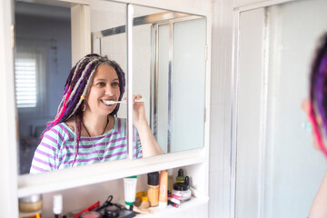 Smiling mature woman with colorful dreadlocks and curvy body brushing teeth reflected in bathroom mirror