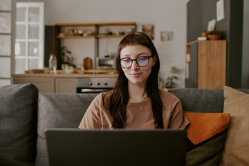 Portrait of young adult Caucasian woman sitting on sofa using laptop for online shopping smiling slightly in modern home interior
