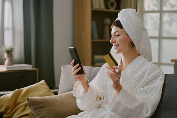 Caucasian young adult woman sitting on sofa smiling while holding smartphone and credit card, wearing bathrobe and towel on head