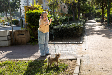 Mature woman with dreadlocks and curvy body walking small dog on leash in sunny city park
