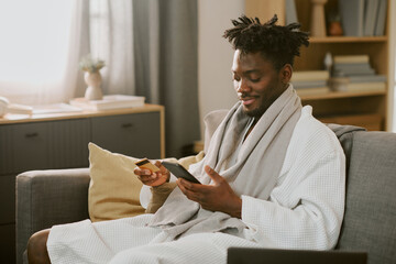 Young adult Black man sitting on sofa holding credit card and smartphone, smiling while making online purchase, engaging in online shopping activity 
