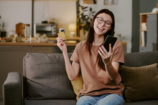 Caucasian young adult woman sitting on sofa holding smartphone and credit card smiling while making online purchase in home setting - Powered by Adobe