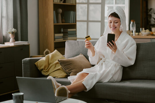 Caucasian young adult woman sitting on sofa wearing bathrobe holding credit card and smartphone smiling while making online purchase with laptop in living room - Powered by Adobe