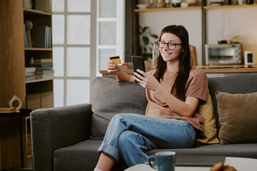 Obraz premium Caucasian young adult woman sitting on sofa holding credit card and smartphone smiling while making online purchase in home setting engaging in digital shopping activity