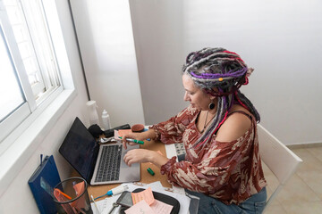 Focused mature woman with colorful dreadlocks and curvy body working on laptop at messy desk