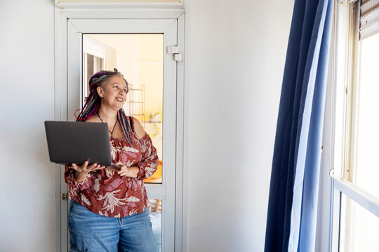 Cheerful mature woman with colorful dreadlocks and curvy body holding laptop and smiling by window - Powered by Adobe