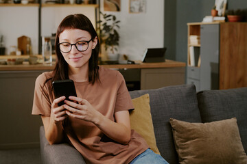 Young adult Caucasian woman sitting on sofa using smartphone for online shopping smiling slightly looking at screen modern home interior visible in background