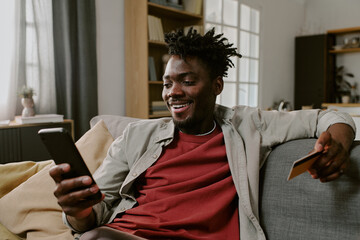 Young adult Black man sitting on sofa holding smartphone in one hand and credit card in other hand smiling while making online shopping transaction 