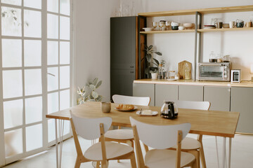 Modern kitchen interior featuring wooden dining table with four chairs, coffee pot, croissant, cup, and plate arranged on tabletop, sunlight streaming through glass doors