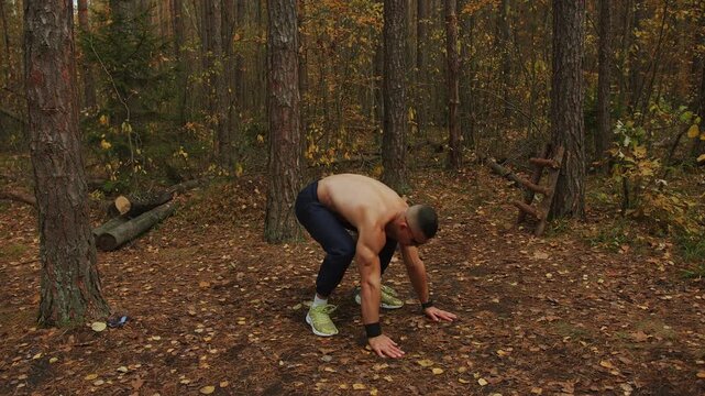 Young muscular man without shirt and wearing sunglasses performs burpee exercise in glade in an autumn forest.