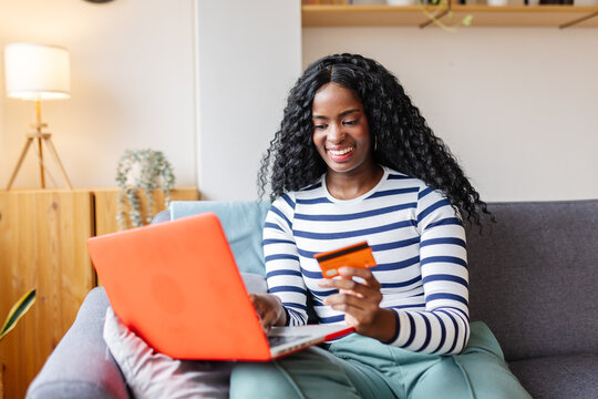 Happy Latin American woman using laptop paying online by credit card sitting on sofa. Joyful black girl shopping online. Female customer making internet payment sitting on sofa at home