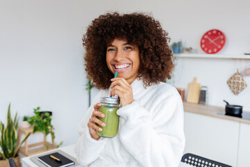 Portrait of young mixed race woman drinking healthy green juice in the morning standing in her kitchen at home. Pretty millennial multiracial girl enjoying bio smoothie shake