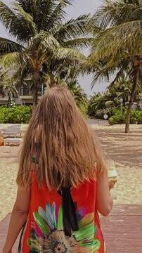 Rear view: a woman in a bright dress walks along a wooden pier to the beach, cocktail in hand; vertical video.
