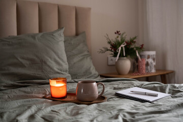 Cozy bedroom scene with a lit candle, warm mug, and notebook on a wooden tray placed on a bed with soft green bedding.